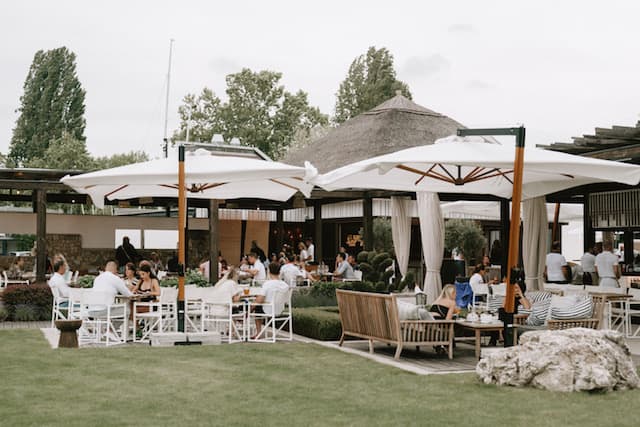 People dining at an outdoor patio with large white umbrellas and a thatched-roof building.