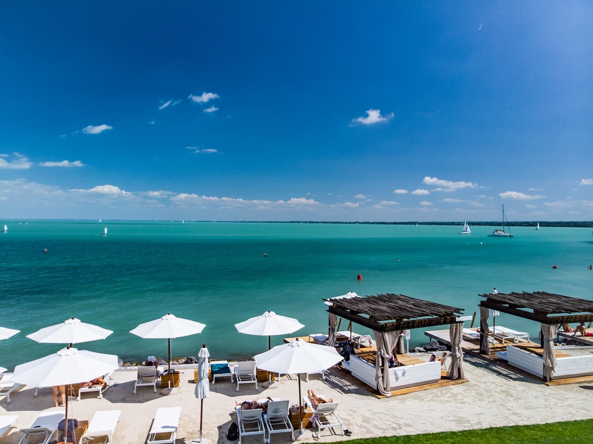 White umbrellas and cabanas line a sandy beach overlooking turquoise water with distant sailboats.