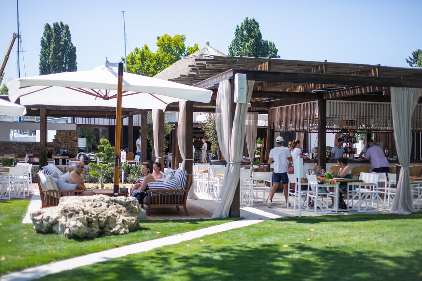 People relaxing at a sunny outdoor restaurant with wooden pergolas and large white umbrellas.