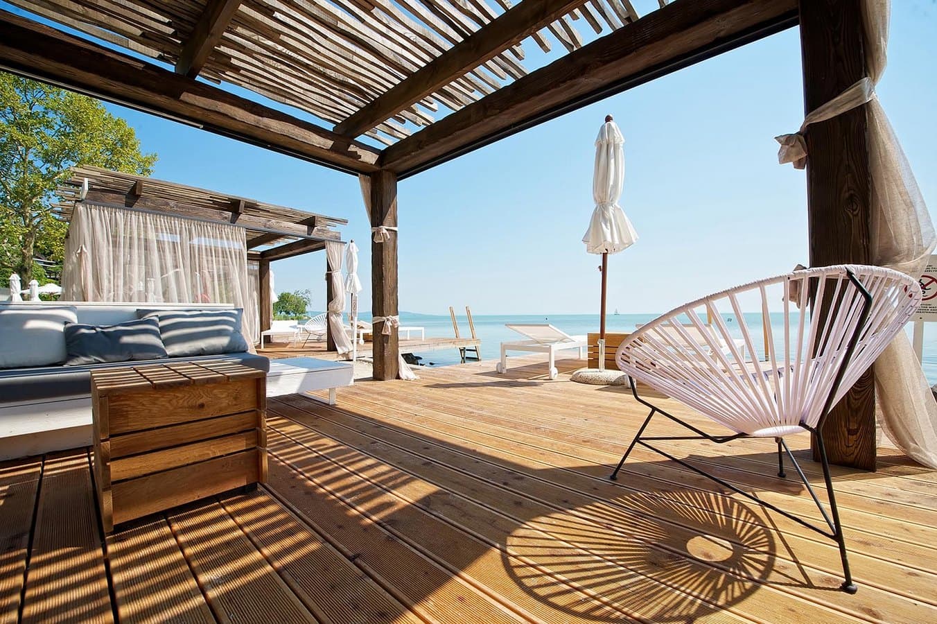 Luxurious wooden deck with white lounge furniture and a pergola overlooking a calm blue lake.