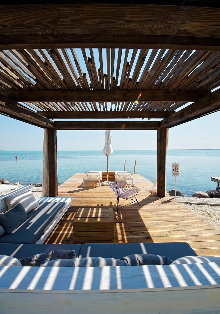 Slatted wooden pergola casts shadows on a deck with lounge seating overlooking calm blue water.