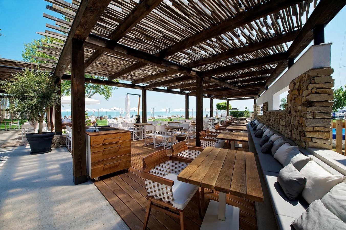 Rustic beachfront patio with wooden tables and chairs under a pergola roof overlooking the sea.
