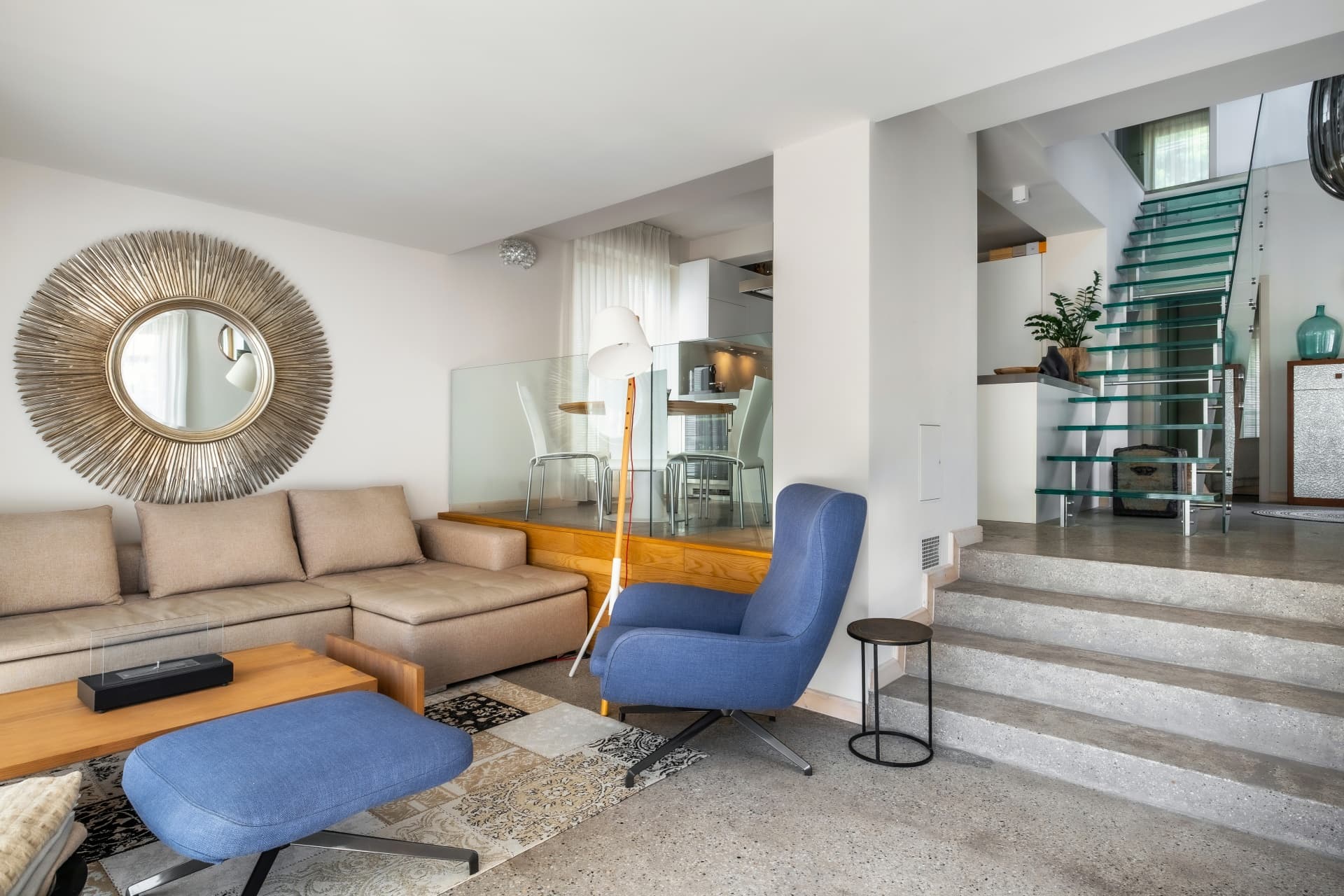 Modern living room with a beige sofa, blue armchair, sunburst mirror, and concrete stairs.