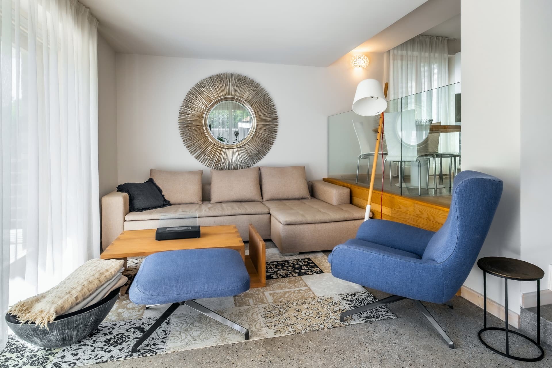 Modern living room with a beige sectional sofa, blue armchair, and a large sunburst mirror.