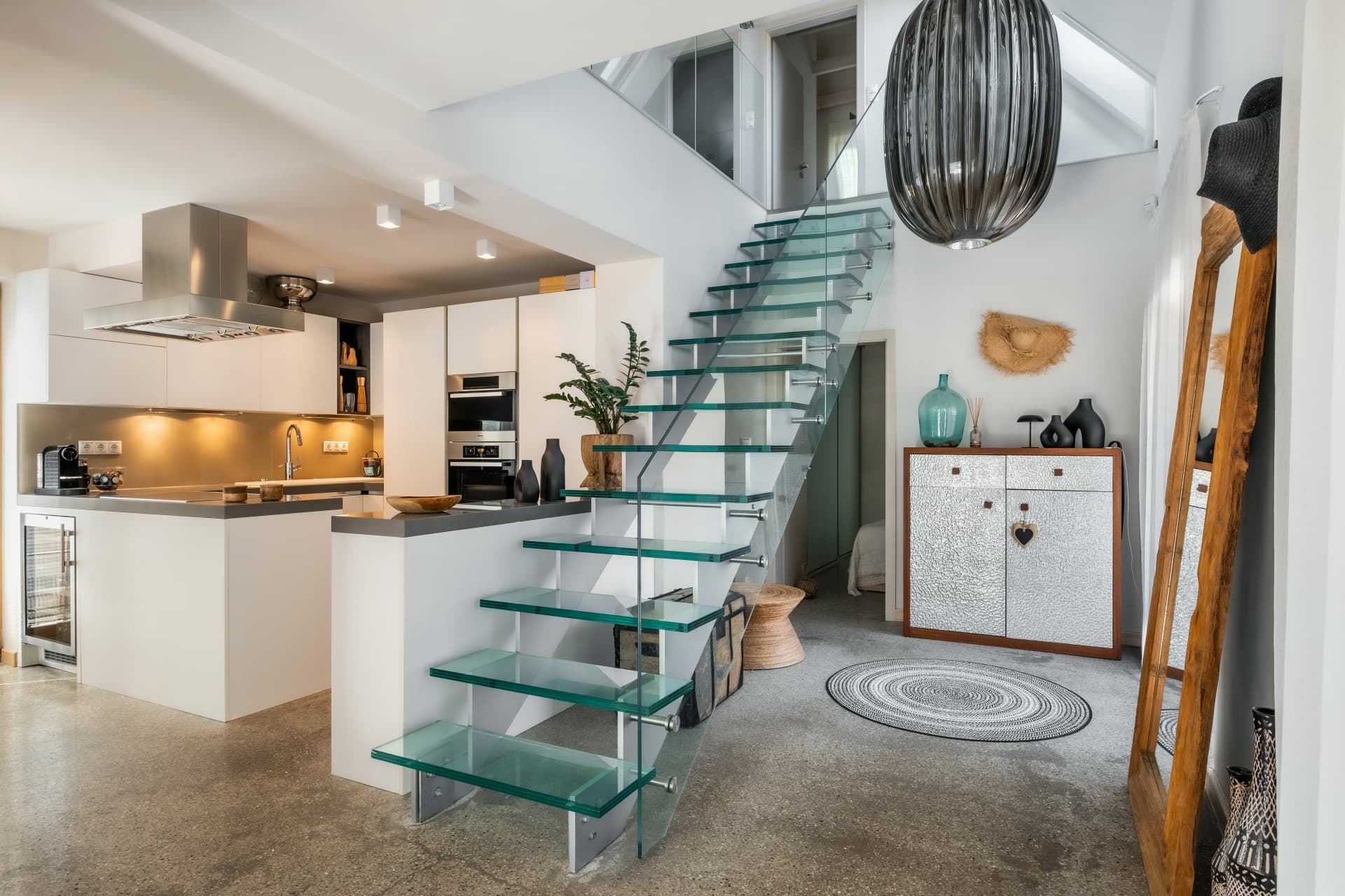 Modern open-plan home featuring a floating glass staircase, white kitchen, and polished concrete floors.
