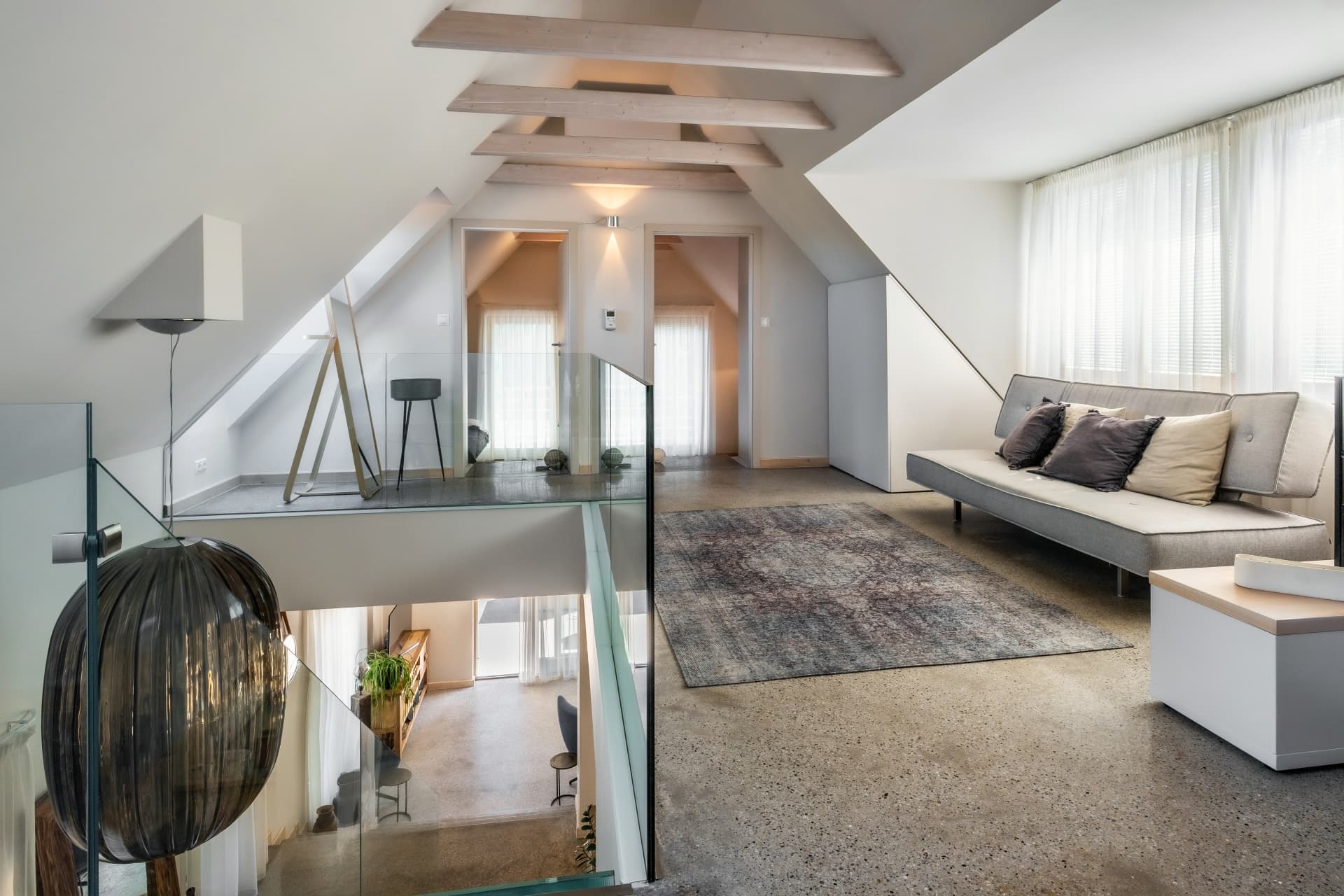 Modern attic living area with slanted ceilings, wooden beams, grey sofa, and glass railings.
