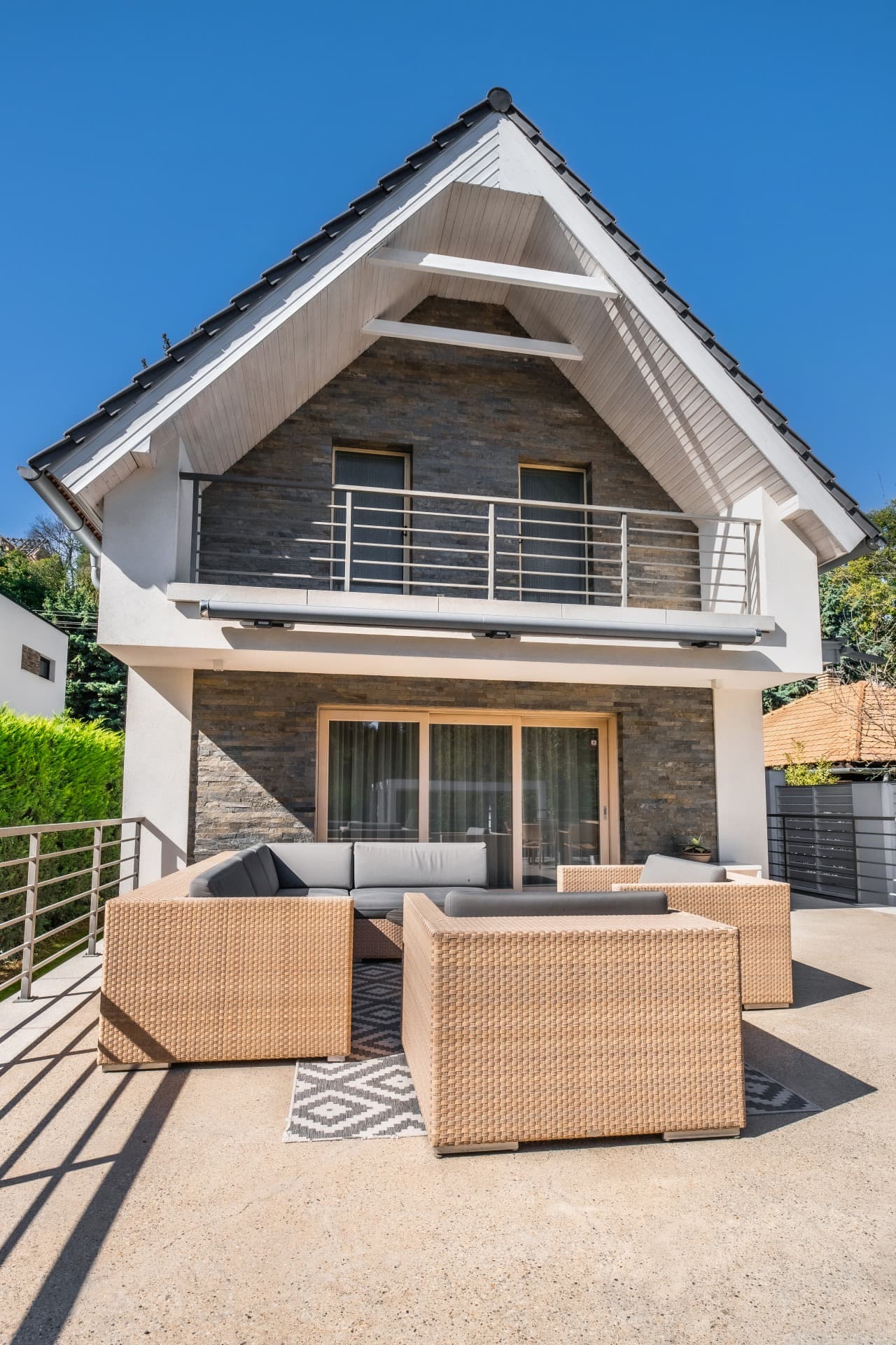 Modern house with stone facade, steep roof, and patio featuring wicker outdoor furniture.