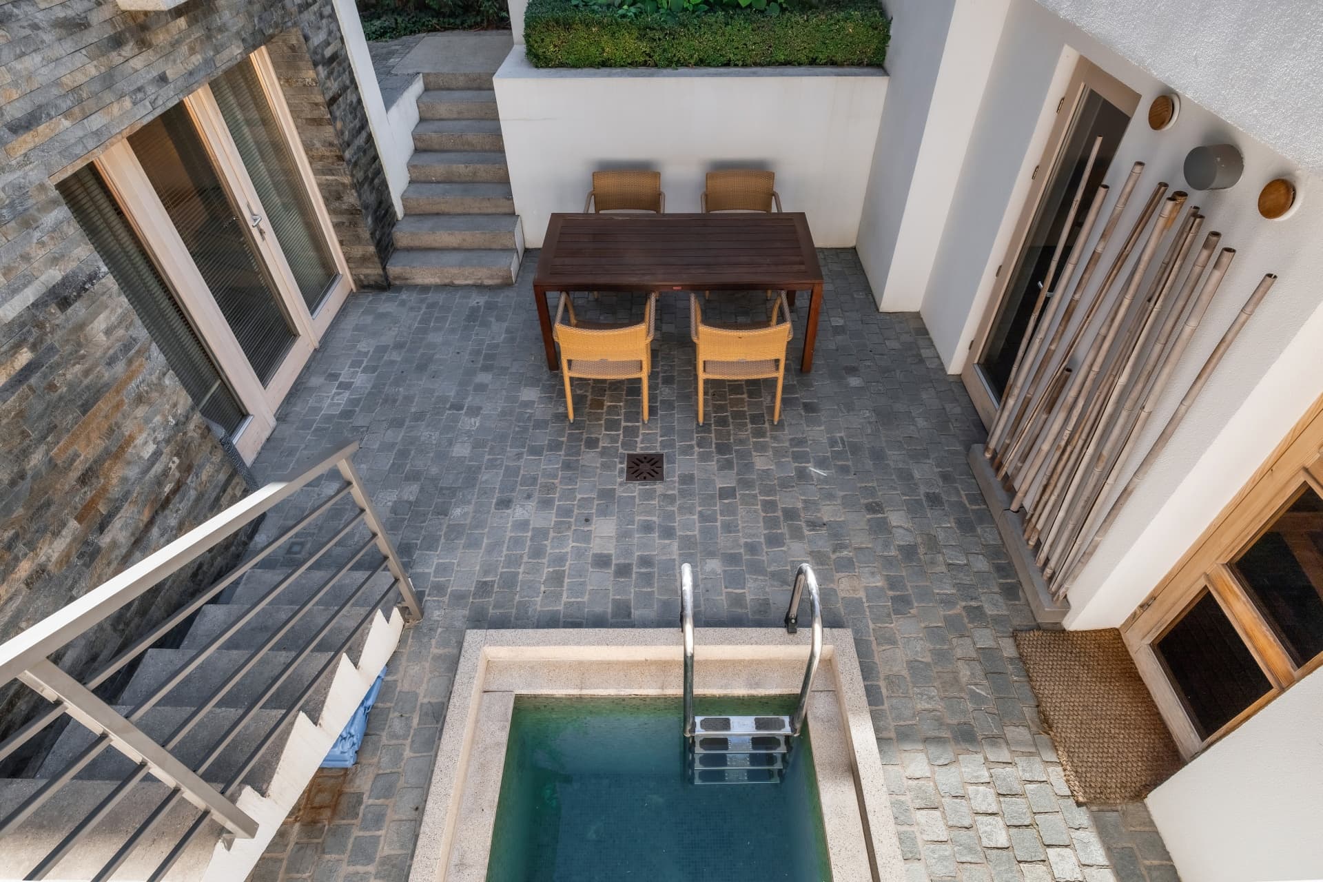 Modern courtyard featuring a small plunge pool, wooden dining table, and grey cobblestone paving.