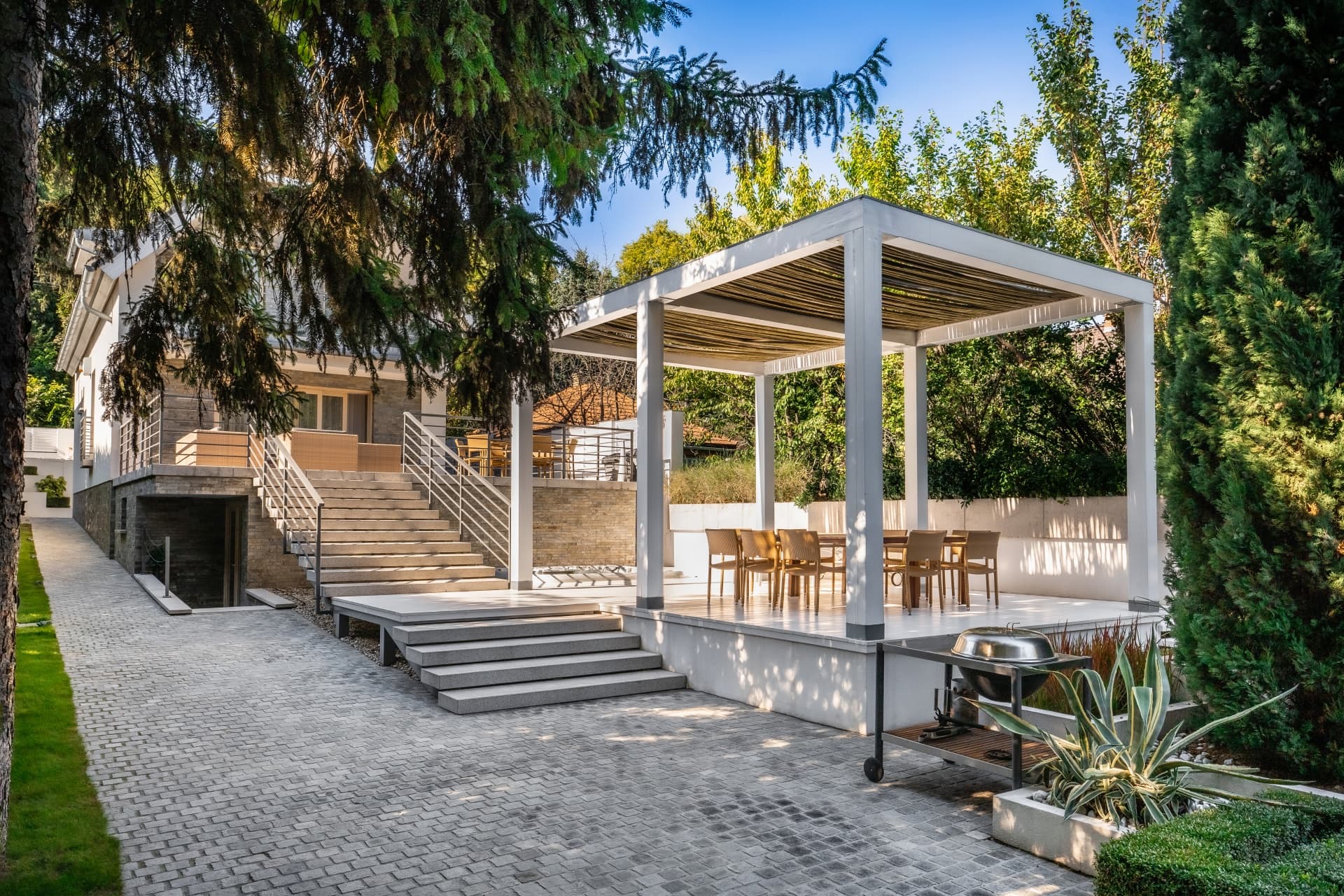 Modern white pergola with dining furniture on a paved patio beside a stone house.